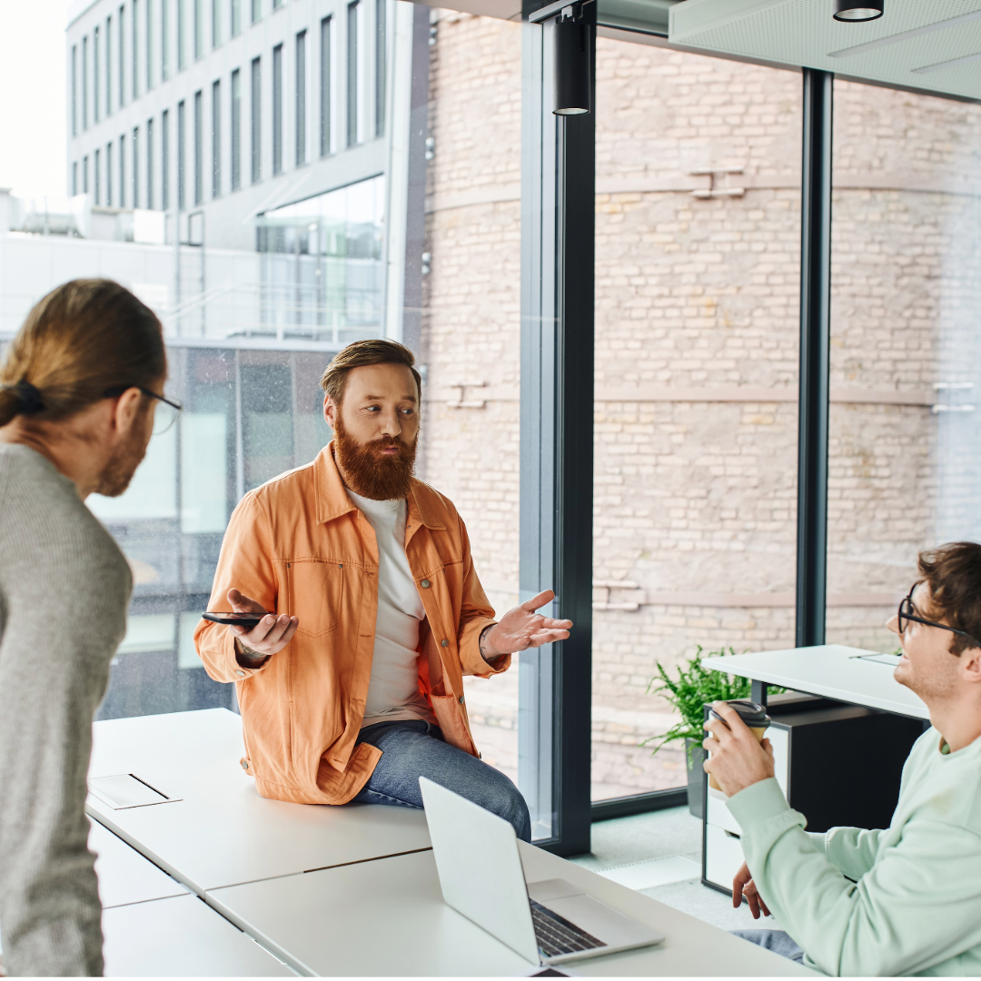 A man sits of the edge of a desk, explaining something to two other people.