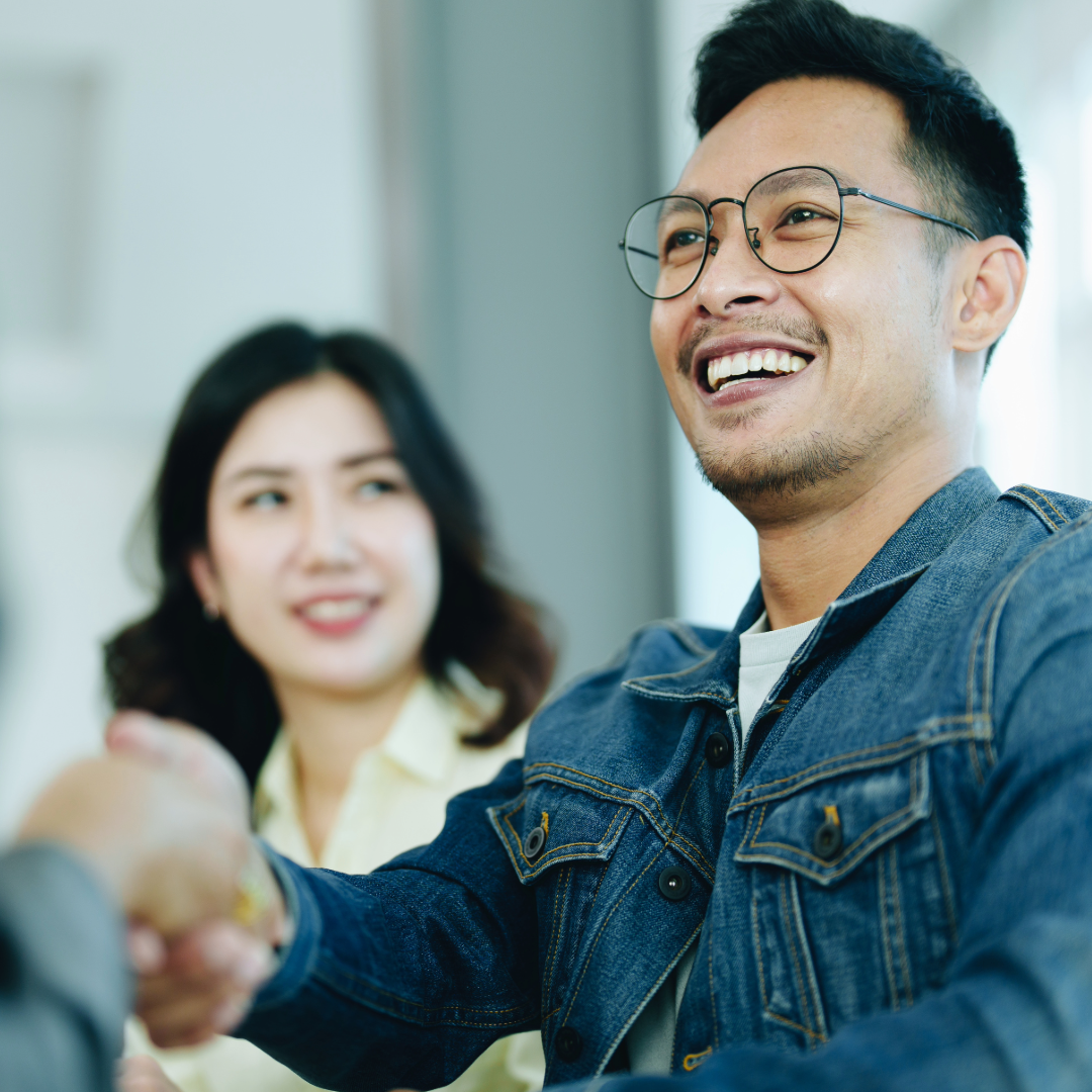 A man shakes hands across a table, watched by another smiling figure.